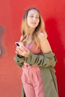 Sosyal medya için cep telefonunu canlı kırmızı bir duvara karşı kullanan, sonbahar atmosferinin tadını çıkaran ve moda modasını gözler önüne seren genç bir stil sahibi.