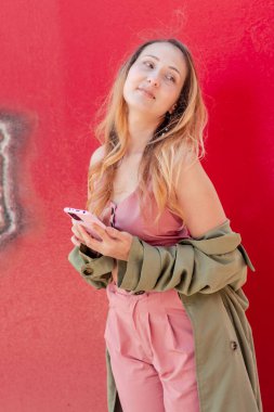 Genç stil sahibi, canlı kırmızı bir duvara karşı poz verirken sosyal medya takipçileri için ilgi çekici bir içerik yaratan bir cep telefonu kullanıyor.