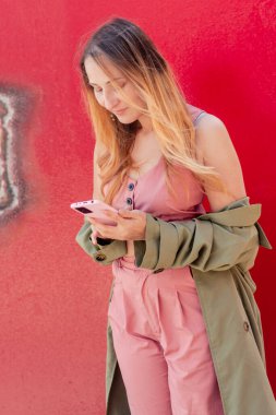 Sosyal medyada akıllı telefonuyla gezinen, canlı kırmızı bir duvara yaslanan, şehir merkezindeki mobil teknolojinin rahatlığından keyif alan, modaya uyan genç bir kadın.