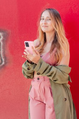 Genç kadın nüfuzlu kişi, canlı kırmızı bir duvara karşı duran sosyal medya takipçilerinin ilgisini çekmek için akıllı telefonunu kullanıyor.