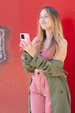 Şık bir genç kadın pembe akıllı telefonu canlı bir kırmızı duvara karşı kullanıyordu, kentsel tarzı ve dijital eğilimleri somutlaştırıyordu.