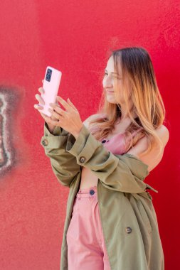 Pembe ve yeşil elbiseli genç kadın nüfuzlu kişi, canlı kırmızı bir duvara karşı durarak sosyal medya için içerik yaratmak için akıllı telefonunu kullanıyor.
