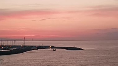 Pembe ve turuncu gökyüzü, sakin bir denizin üzerinde, limandan ayrılan küçük bir balıkçı teknesiyle, sakin ve huzurlu bir manzara yaratarak, açık sulara doğru yelken açıyor.