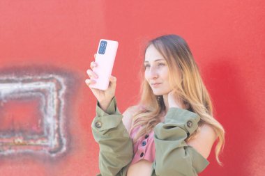 Genç bir kadın pembe bir akıllı telefonla canlı kırmızı bir duvara karşı selfie çekiyor, kendini ifade etmeyi ve modern sosyal medya bağlantısının özünü kucaklıyor.