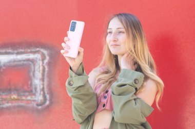 Genç bir kadın kırmızı duvarın önünde pembe akıllı telefonuyla selfie çekiyor, sosyal medya için içerik yaratıyor, tek eliyle boynuna dokunuyor, modaya uygun kıyafetler giyiyor.