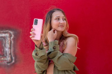 Kırmızı bir duvarın yanında poz verirken pembe akıllı telefonunu göstererek takipçileri için canlı bir sonbahar sezonunda ilgi çekici bir içerik yaratıyor.