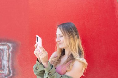 Sosyal medya için canlı içerikler yakalayan, akıllı telefonuyla çarpıcı bir kırmızı duvara karşı fotoğraf çeken, güneşli bir sonbahar gününün sıcaklığından zevk alan bir tetikçi.