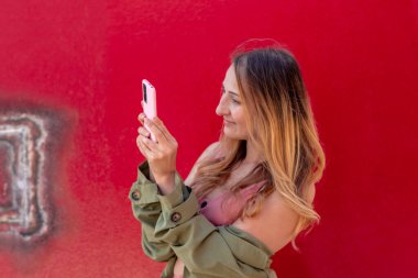 Genç sarışın kadın turist, güneşli bir yaz gününde, kırmızı bir duvarın önünde, paslı bir boyayla pembe akıllı telefonuyla fotoğraf çekiyor.