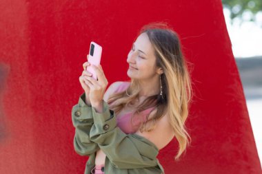 Pembe akıllı telefonuyla fotoğraf çeken mutlu genç bir kadın, hareketli bir şehir ortamında bir anlık keyfi yaşıyor çarpıcı kırmızı arka planda, sonbaharda yaptığı seyahatler sırasında anılarını yakalıyor.