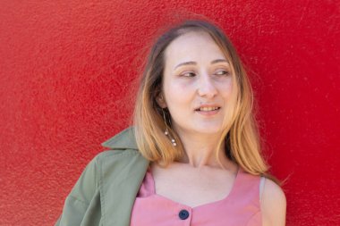 Açık renk elbiseli ve şık küpeli sarışın kadın canlı kırmızı bir duvara karşı poz veriyor. İnce bir gülümseme, ışık saçan bir güven ve sıradan bir güzellikle başka tarafa bakıyor.