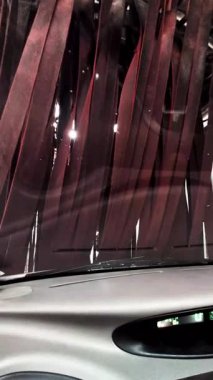 Otomatik araba yıkama sırasında ön camı temizleyen canlı kırmızı fırçalar, içerideki aracın sabunlu yıkama hareketiyle iç perspektifi yakalıyor.