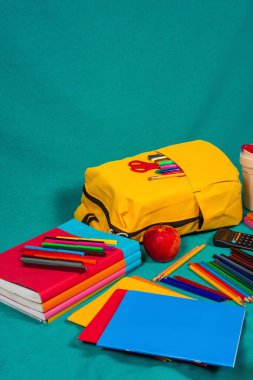 Not defteri, kalem ve kırmızı elma gibi renkli okul gereçleri turkuaz arka planda sarı bir sırt çantasının etrafını sararak okul yılına hazırlık sinyalleri veriyor.