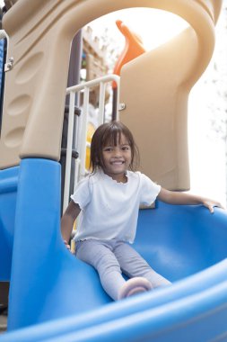 Genç bir kız gökyüzü mavisi kaydırağında oturuyor, gülümsüyor ve el sallıyor. Eğlence ve heyecan kavramı, kız çocuk parkında eğlenirken.