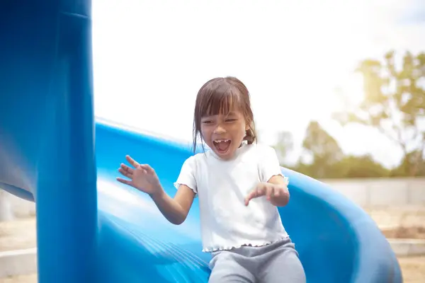 stock image A young girl is sitting on a sky blue slide, smiling and waving. Concept of joy and excitement, as the girl is enjoying her time at the playground