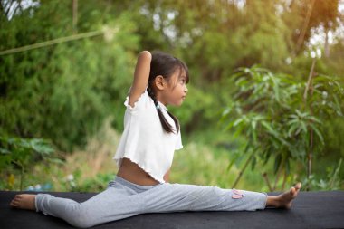 Küçük bir kız bahçede yoga yapıyor, doğanın ferahlatıcı kucaklamalarıyla çevrili, hareketleri kuşlar ve hışırdayan yapraklarla uyumlu, gün ışığı ağaçların arasından süzülürken.