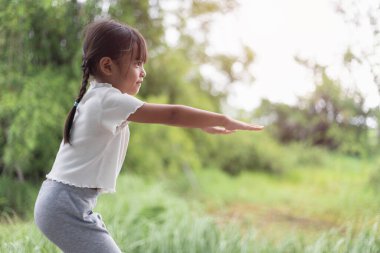 Küçük bir kız bahçede yoga yapıyor, doğanın ferahlatıcı kucaklamalarıyla çevrili, hareketleri kuşlar ve hışırdayan yapraklarla uyumlu, gün ışığı ağaçların arasından süzülürken.