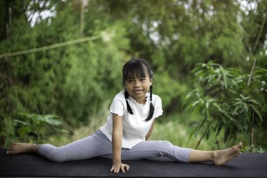 Küçük bir kız bahçede yoga yapıyor, doğanın ferahlatıcı kucaklamalarıyla çevrili, hareketleri kuşlar ve hışırdayan yapraklarla uyumlu, gün ışığı ağaçların arasından süzülürken.