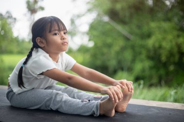 Küçük bir kız bahçede yoga yapıyor, doğanın ferahlatıcı kucaklamalarıyla çevrili, hareketleri kuşlar ve hışırdayan yapraklarla uyumlu, gün ışığı ağaçların arasından süzülürken.