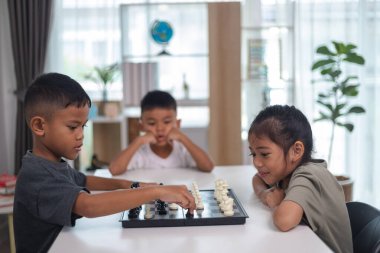 İki çocuk satranç oynuyor. Biri erkek, diğeri de bir kız. Önlerinde bir satranç tahtasıyla bir masada oturuyorlar.