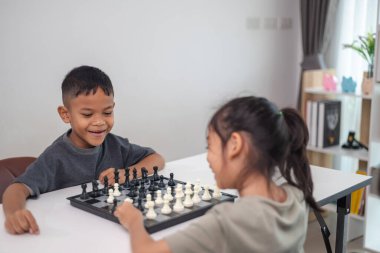 İki çocuk satranç oynuyor. Biri erkek, diğeri de bir kız. Önlerinde bir satranç tahtasıyla bir masada oturuyorlar.