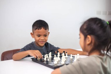 İki çocuk satranç oynuyor. Biri erkek, diğeri de bir kız. Önlerinde bir satranç tahtasıyla bir masada oturuyorlar.