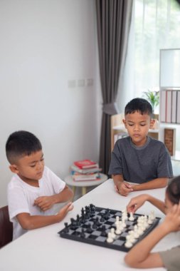 İki çocuk satranç oynuyor. Biri erkek, diğeri de bir kız. Önlerinde bir satranç tahtasıyla bir masada oturuyorlar.