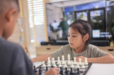 İki çocuk satranç oynuyor. Biri erkek, diğeri de bir kız. Önlerinde bir satranç tahtasıyla bir masada oturuyorlar.