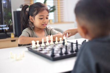 İki çocuk satranç oynuyor. Biri erkek, diğeri de bir kız. Önlerinde bir satranç tahtasıyla bir masada oturuyorlar.