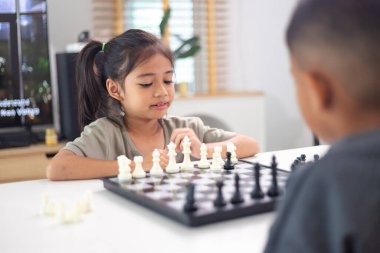 İki çocuk satranç oynuyor. Biri erkek, diğeri de bir kız. Önlerinde bir satranç tahtasıyla bir masada oturuyorlar.