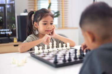İki çocuk satranç oynuyor. Biri erkek, diğeri de bir kız. Önlerinde bir satranç tahtasıyla bir masada oturuyorlar.