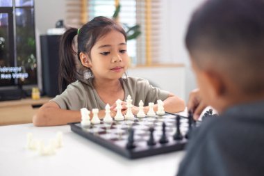 İki çocuk satranç oynuyor. Biri erkek, diğeri de bir kız. Önlerinde bir satranç tahtasıyla bir masada oturuyorlar.