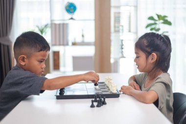 İki çocuk satranç oynuyor. Biri erkek, diğeri de bir kız. Önlerinde bir satranç tahtasıyla bir masada oturuyorlar.