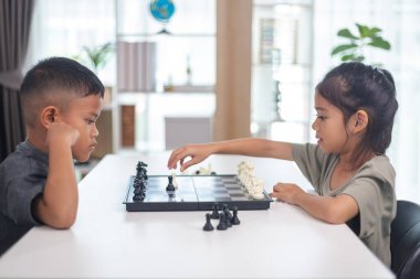 İki çocuk satranç oynuyor. Biri erkek, diğeri de bir kız. Önlerinde bir satranç tahtasıyla bir masada oturuyorlar.