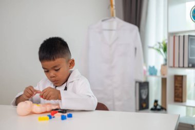 Genç bir çocuk doktorun ofisinde bir bebekle oynuyor. Oda steteskop ve oyuncak doktor seti de dahil olmak üzere tıbbi ekipmanlarla dolu. Çocuk beyaz bir laboratuvar önlüğü giyiyor.