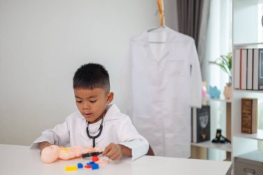 Genç bir çocuk doktorun ofisinde bir bebekle oynuyor. Oda steteskop ve oyuncak doktor seti de dahil olmak üzere tıbbi ekipmanlarla dolu. Çocuk beyaz bir laboratuvar önlüğü giyiyor.