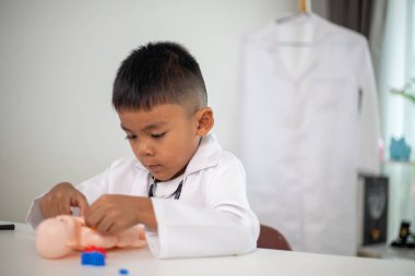Genç bir çocuk doktorun ofisinde bir bebekle oynuyor. Oda steteskop ve oyuncak doktor seti de dahil olmak üzere tıbbi ekipmanlarla dolu. Çocuk beyaz bir laboratuvar önlüğü giyiyor.