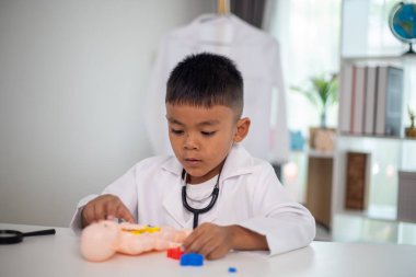 Genç bir çocuk doktorun ofisinde bir bebekle oynuyor. Oda steteskop ve oyuncak doktor seti de dahil olmak üzere tıbbi ekipmanlarla dolu. Çocuk beyaz bir laboratuvar önlüğü giyiyor.
