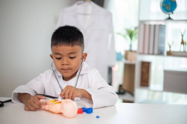 Genç bir çocuk doktorun ofisinde bir bebekle oynuyor. Oda steteskop ve oyuncak doktor seti de dahil olmak üzere tıbbi ekipmanlarla dolu. Çocuk beyaz bir laboratuvar önlüğü giyiyor.