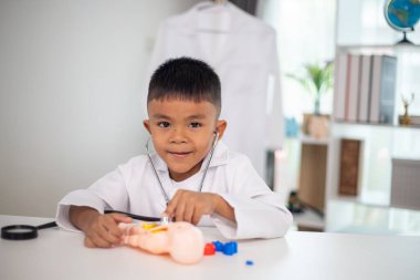 Genç bir çocuk doktorun ofisinde bir bebekle oynuyor. Oda steteskop ve oyuncak doktor seti de dahil olmak üzere tıbbi ekipmanlarla dolu. Çocuk beyaz bir laboratuvar önlüğü giyiyor.