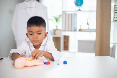 Genç bir çocuk doktorun ofisinde bir bebekle oynuyor. Oda steteskop ve oyuncak doktor seti de dahil olmak üzere tıbbi ekipmanlarla dolu. Çocuk beyaz bir laboratuvar önlüğü giyiyor.