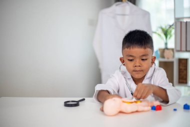 Genç bir çocuk doktorun ofisinde bir bebekle oynuyor. Oda steteskop ve oyuncak doktor seti de dahil olmak üzere tıbbi ekipmanlarla dolu. Çocuk beyaz bir laboratuvar önlüğü giyiyor.