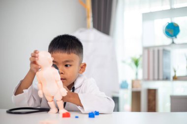 Genç bir çocuk doktorun ofisinde bir bebekle oynuyor. Oda steteskop ve oyuncak doktor seti de dahil olmak üzere tıbbi ekipmanlarla dolu. Çocuk beyaz bir laboratuvar önlüğü giyiyor.