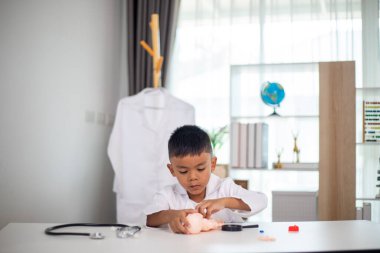 Genç bir çocuk doktorun ofisinde bir bebekle oynuyor. Oda steteskop ve oyuncak doktor seti de dahil olmak üzere tıbbi ekipmanlarla dolu. Çocuk beyaz bir laboratuvar önlüğü giyiyor.