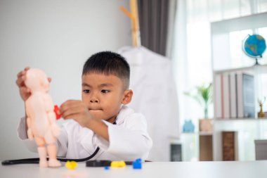 Genç bir çocuk doktorun ofisinde bir bebekle oynuyor. Oda steteskop ve oyuncak doktor seti de dahil olmak üzere tıbbi ekipmanlarla dolu. Çocuk beyaz bir laboratuvar önlüğü giyiyor.