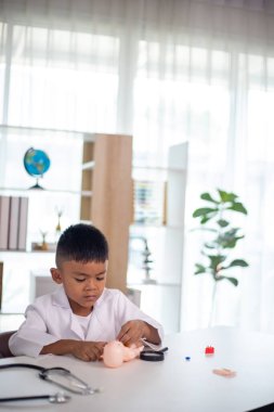 Genç bir çocuk doktorun ofisinde bir bebekle oynuyor. Oda steteskop ve oyuncak doktor seti de dahil olmak üzere tıbbi ekipmanlarla dolu. Çocuk beyaz bir laboratuvar önlüğü giyiyor.