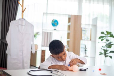 Genç bir çocuk doktorun ofisinde bir bebekle oynuyor. Oda steteskop ve oyuncak doktor seti de dahil olmak üzere tıbbi ekipmanlarla dolu. Çocuk beyaz bir laboratuvar önlüğü giyiyor.
