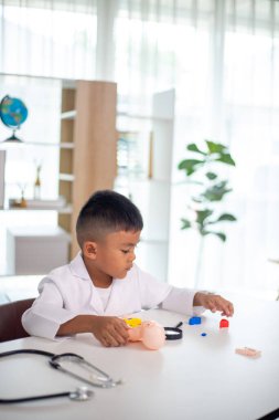 Genç bir çocuk doktorun ofisinde bir bebekle oynuyor. Oda steteskop ve oyuncak doktor seti de dahil olmak üzere tıbbi ekipmanlarla dolu. Çocuk beyaz bir laboratuvar önlüğü giyiyor.