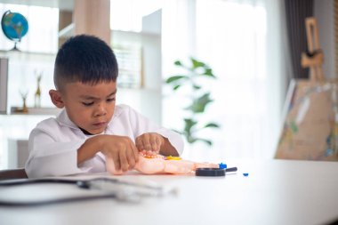 Genç bir çocuk doktorun ofisinde bir bebekle oynuyor. Oda steteskop ve oyuncak doktor seti de dahil olmak üzere tıbbi ekipmanlarla dolu. Çocuk beyaz bir laboratuvar önlüğü giyiyor.
