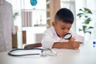 Genç bir çocuk doktorun ofisinde bir bebekle oynuyor. Oda steteskop ve oyuncak doktor seti de dahil olmak üzere tıbbi ekipmanlarla dolu. Çocuk beyaz bir laboratuvar önlüğü giyiyor.