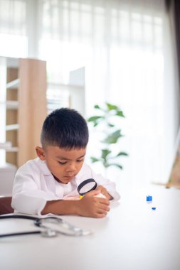 Genç bir çocuk doktorun ofisinde bir bebekle oynuyor. Oda steteskop ve oyuncak doktor seti de dahil olmak üzere tıbbi ekipmanlarla dolu. Çocuk beyaz bir laboratuvar önlüğü giyiyor.